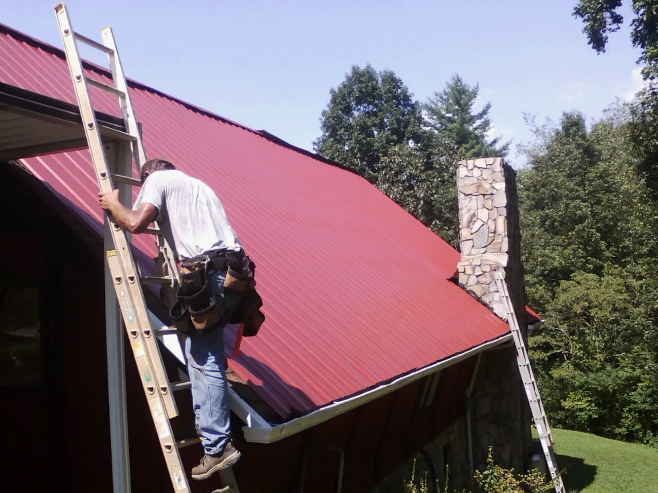 Roofer on a ladder beside a red metal roof and stone chimney — Soffit & Fascia Repair by Roofing Brothers in Longview